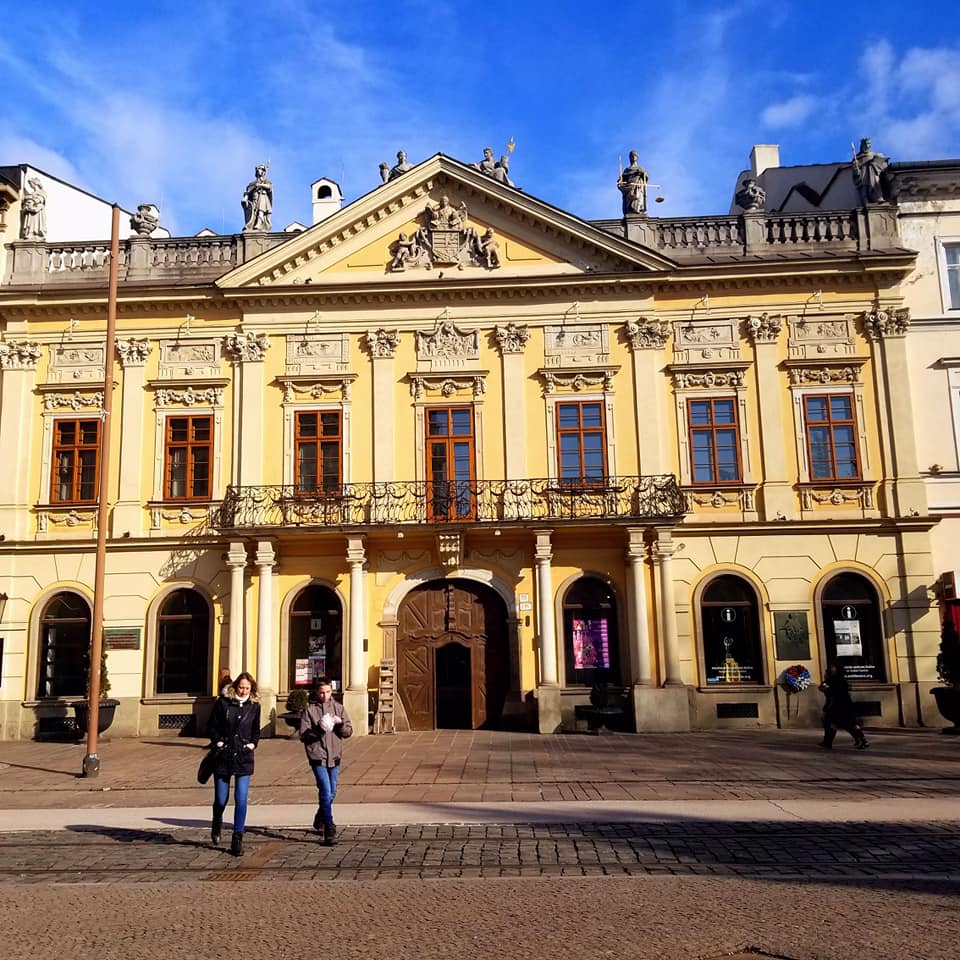 Exterior of a large, pale yellow building in Kosice, Slovakia. There are two women walking away from the building and towards the camera. The skies in the background are blue with a few whispy clouds.