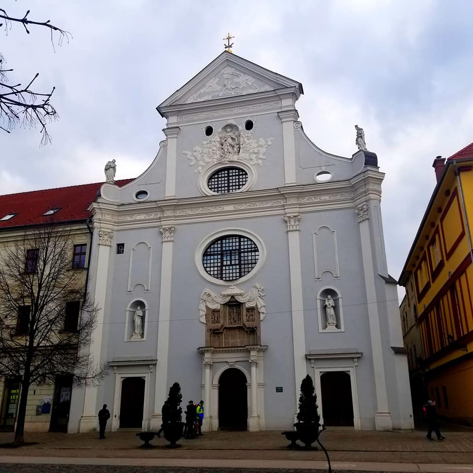 Pale blue church in Kosice Slovakia. The building has white trim, and two circular windows above the main entrance. There are red buildings on either side of the church. The sky is also a pale blue with a few clouds in the background.
