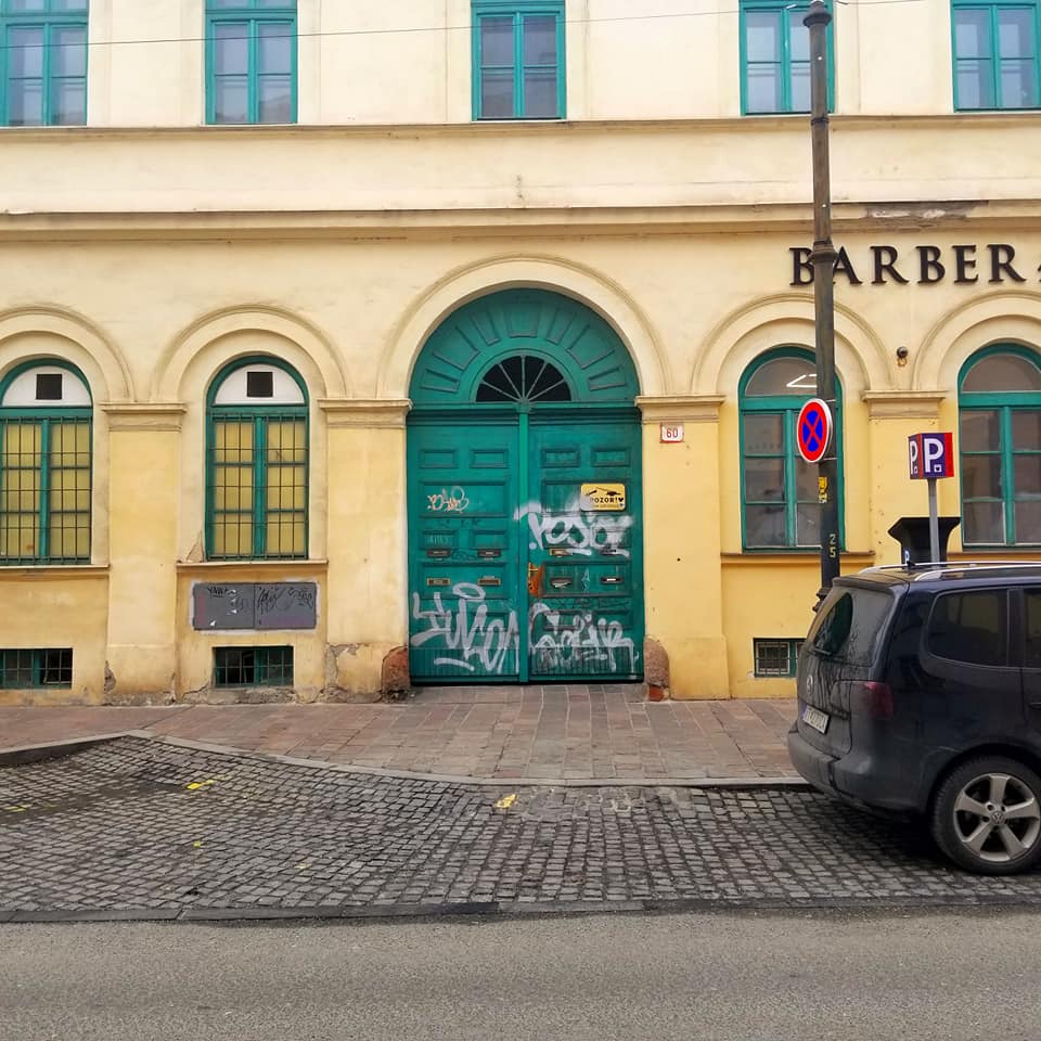 Bright yellow building with teal doors in Kosice, Slovakia. There is white graffiti all over the arced door. The street in front of the building is cobblestone and there is a sign that says 'BARBER' in the upper right hand corner. There is the rear portion of a dark vehicle parked on the street on the right with a street pole behind it.