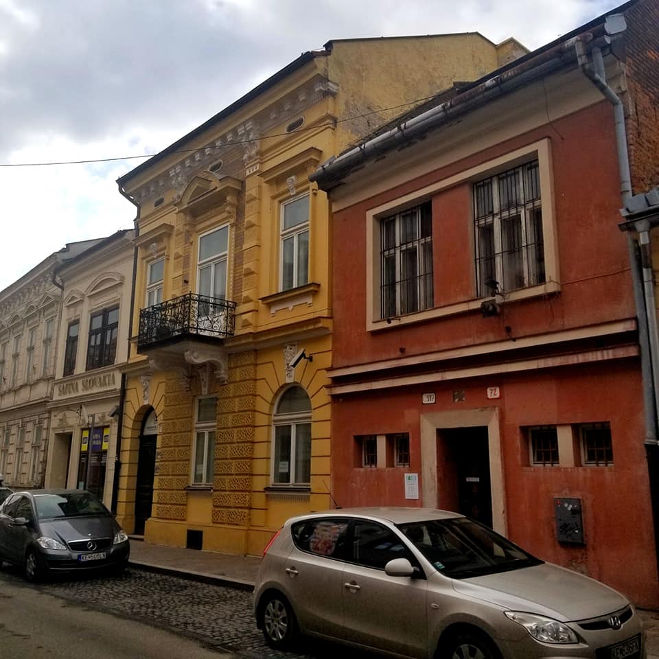 Looking down the street in Kosice Slovakia. There are cars lining the cobblestone road with three buildings visible. The farthest is cream colored, then deep mustard yellow, and then a red building.