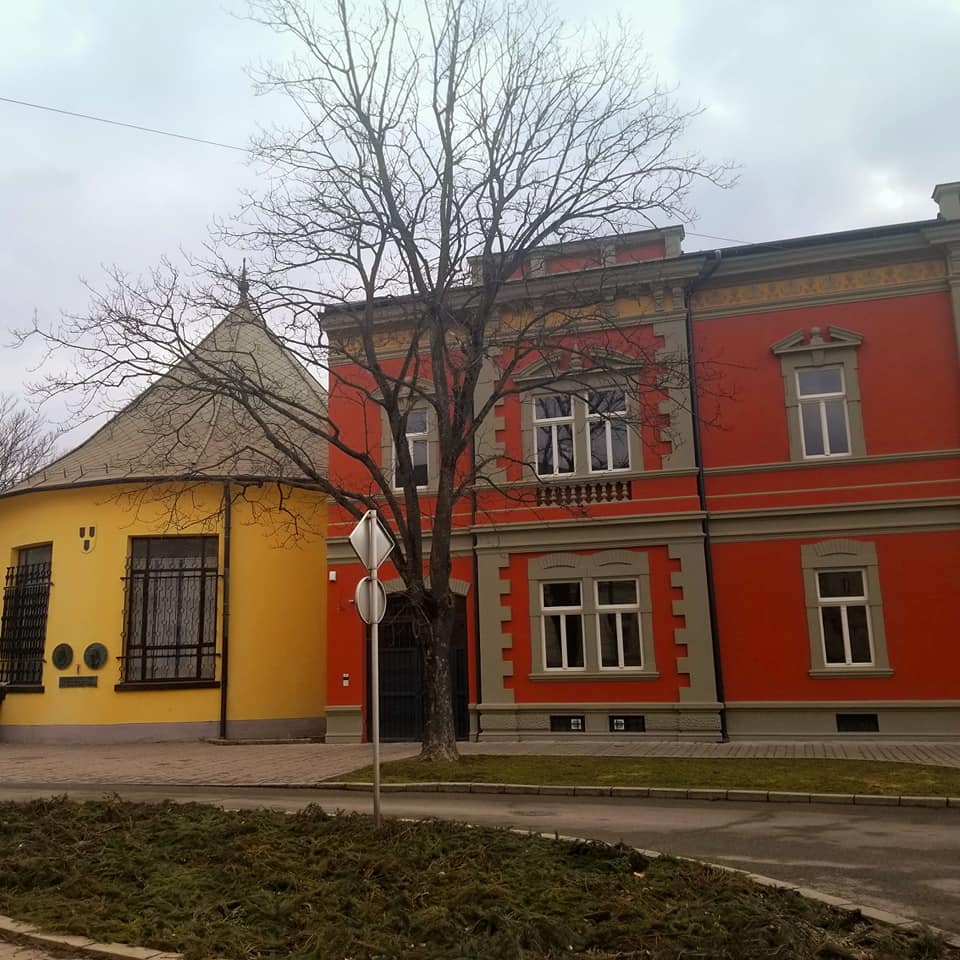 In Kosice, Slovakia: Round yellow building with a conical roof adjacent to a red building with tan trim. In front of the building there is a street sign and a large tree with no leaves. The sky is bleak and dreary looking and the street in front of the buildings are cobblestone.