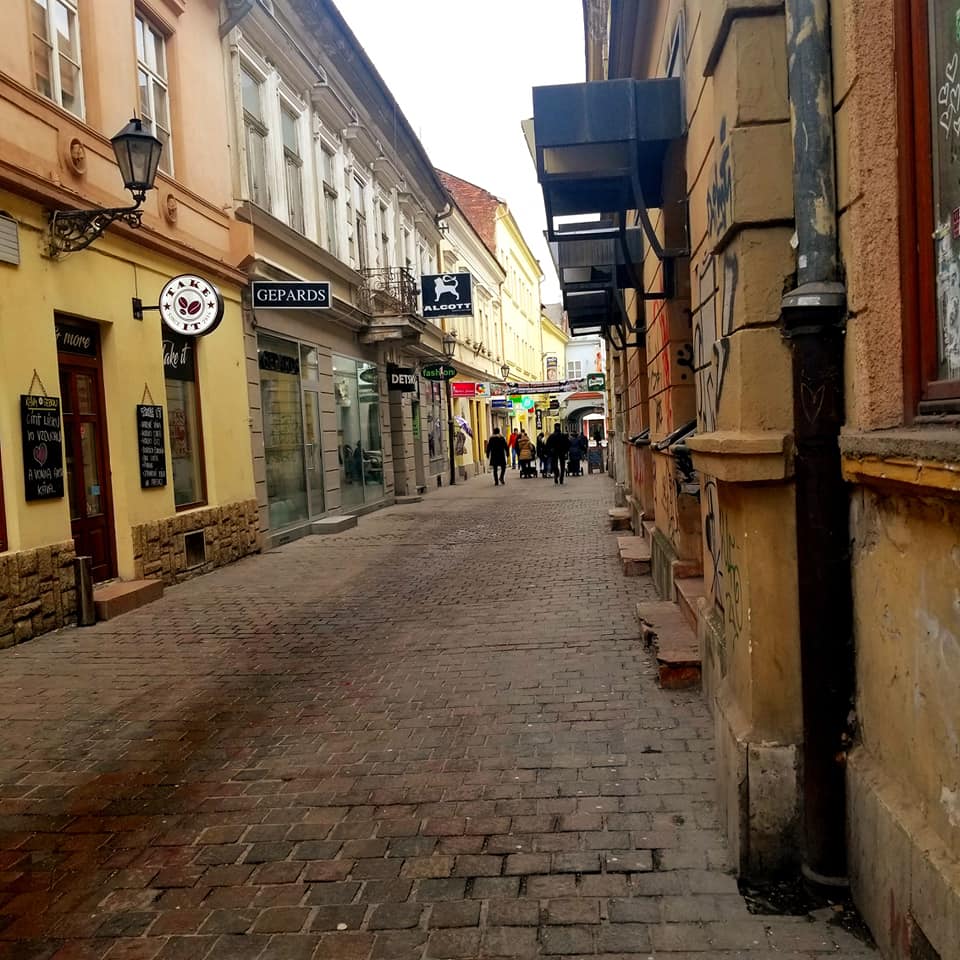 Street in Kosice, Slovakia made of cobblestone. The street winds to the right. The buildings on either side are old, yet preserved, in shades of yellow, ochre, pale orange. There are stores and restaurants lining the streets. In the distance you can see a group of people walking in the street.