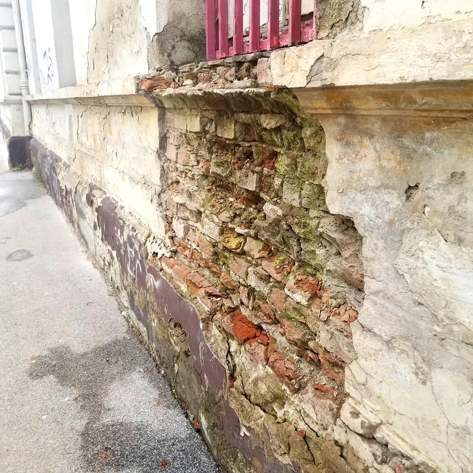 Decaying building in Kosice Slovakia. The building is cream colored stone, and there is a window wiith red vertical bars. In the space beneath the window, the stone is crumbling off of the building revealing bricks underneath.