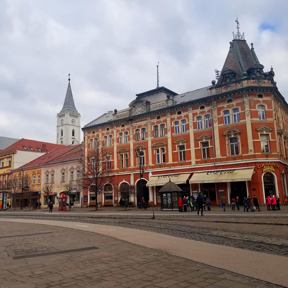 Exterior of a red building in Kosice, Slovakia. The building has cream colored trim and a black roof. There are cream colored awnings in the front of the building. The street in front of the building is made of cobblestone. There are a few people immediately in front of the building.