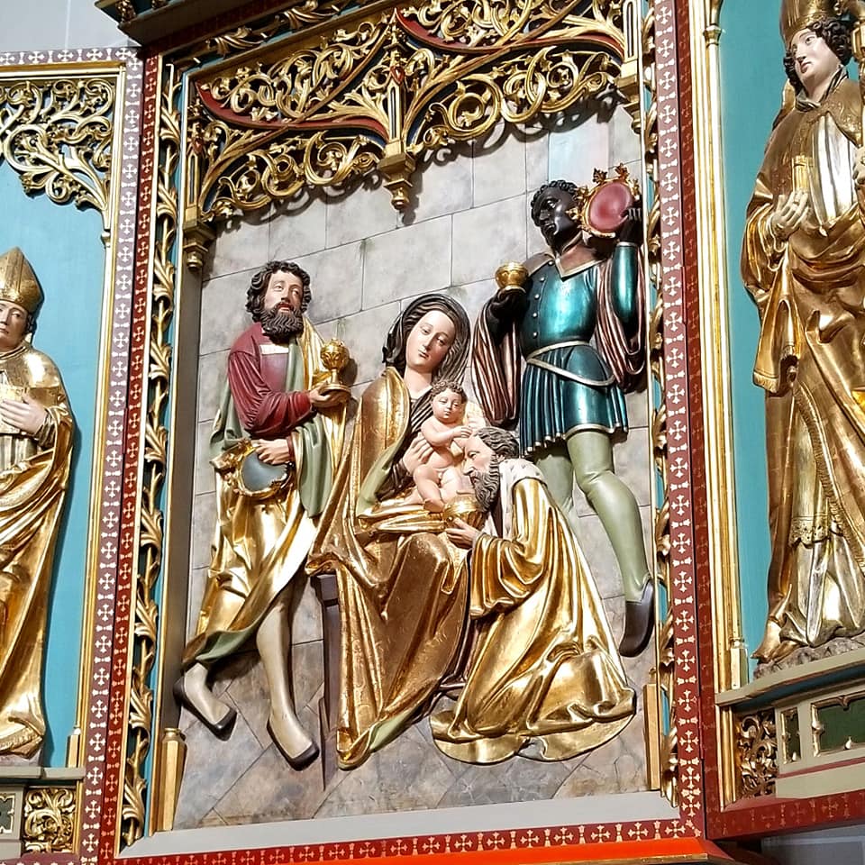 Religious carving at Saint Elisabeth's cathedral in Kosice, Slovakia. There is gold leaf with shades of red and teal. The carving is of one seated woman with someone crying in her lap and two people in the background
