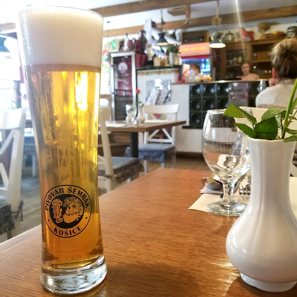 Close-up picture of a tall pale-colored beer with an inch and a half of head in a pilsner glass. Also on the table is an empty water glass and a small vase with a single red rose in it. Blurred in the background is the bar of the restaurant, empty.