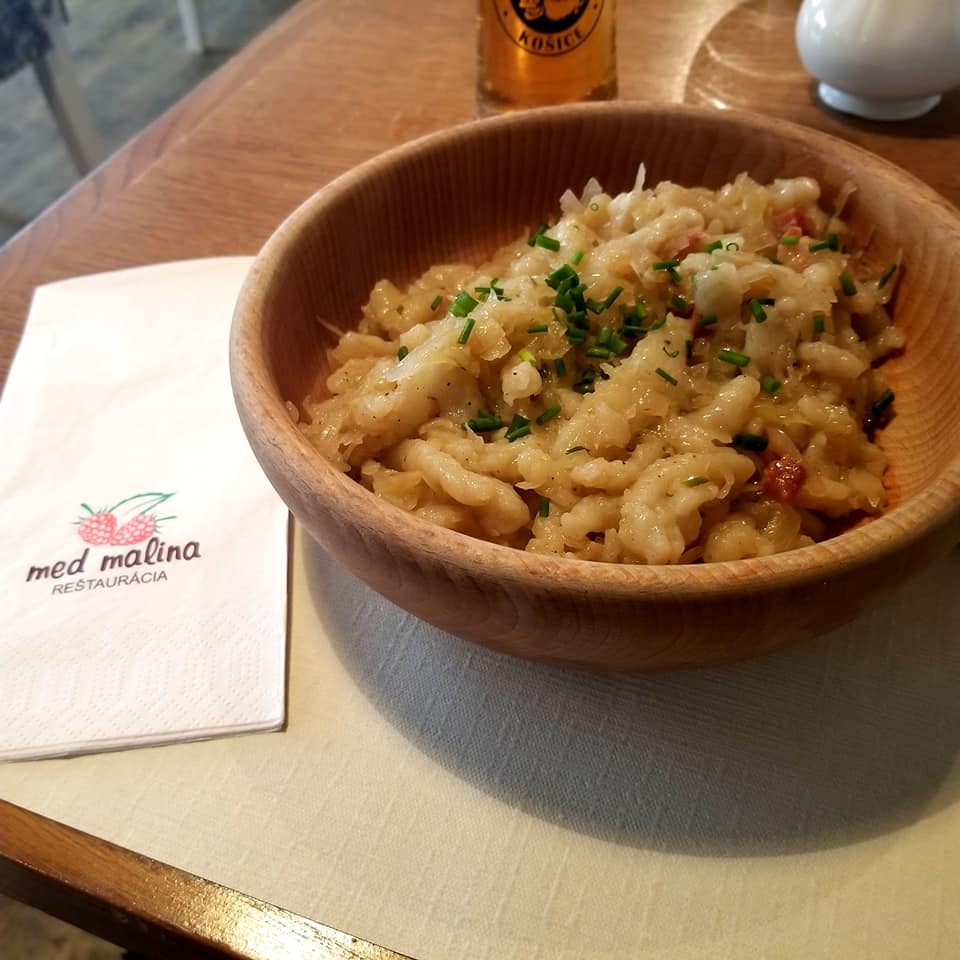 Traditional Eastern Slovak dish of potato dumplings with roasted bacon and sour cabbage served in a bowl. Next to the bowl is a napkin with the 'Med Malina' logo on it with a beer in the background.