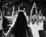 Black and white photograph of a group of people outside practicing yoga.  Their hands are up toward the sky and they are gazing at their hands.  An instructor is standing with her back to the camera, long dark curly hair.  In the background are the people practicing yoga.  There are trees in the background but they are not easy to see because the photo is sharp black and white.