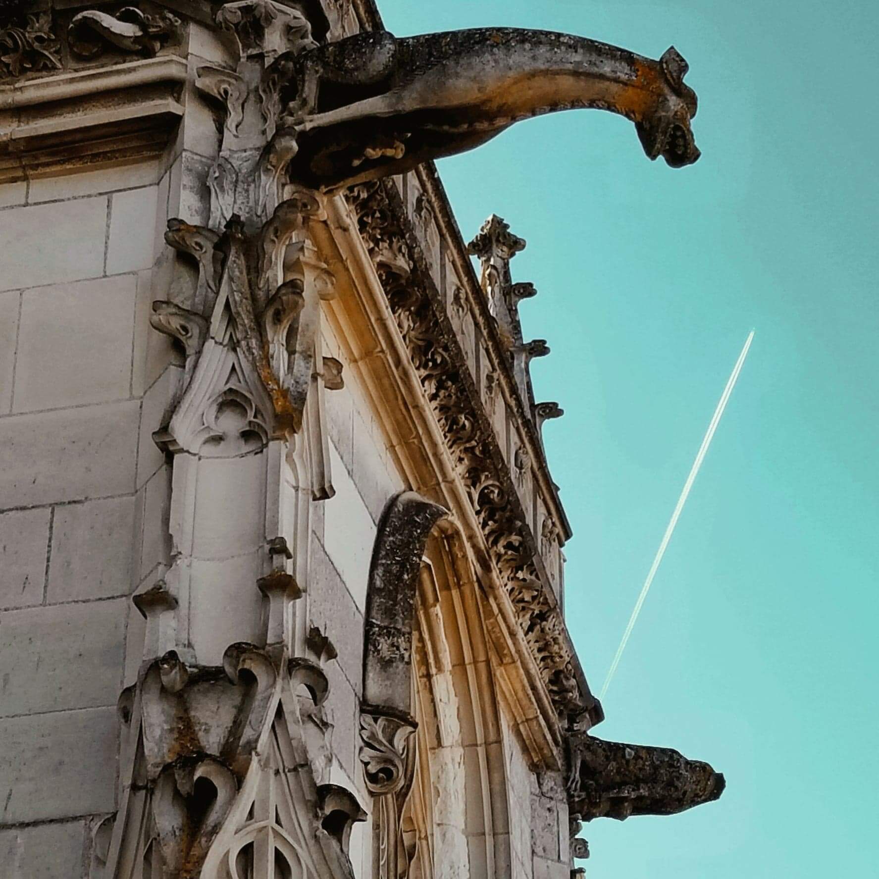 Looking up at the eave of a castle in France with gargoyles looking down, set agains the sky. The building is detailed and old. There are two gargoyles. In the clear blue sky, there is a streak of white from a plane flying by.