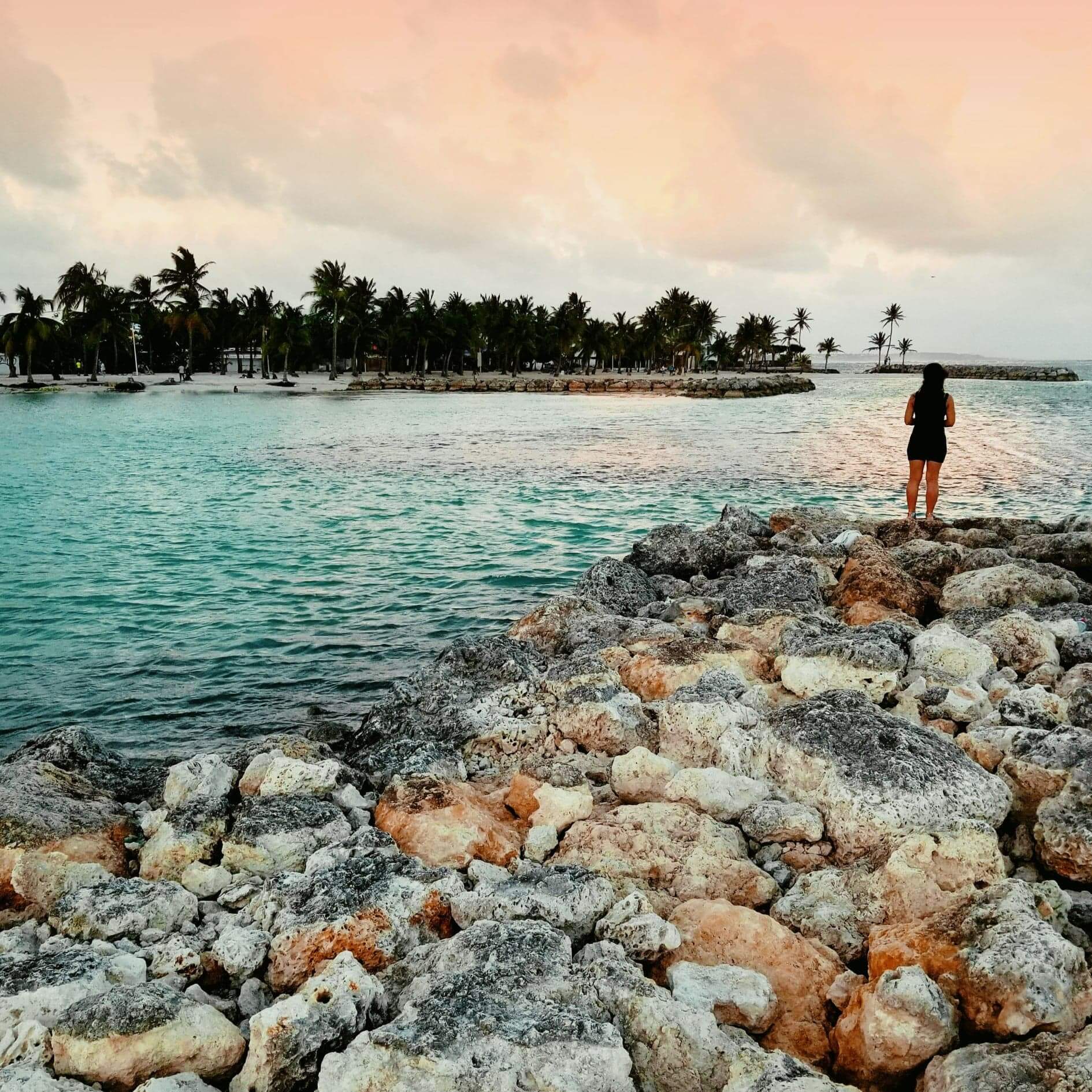 Woman standing at the end of a rocky pier during sunset. The water is turquoise and tropical and the sky is orange-ish pink. In the background there is a small isle with a number of trees. The woman is wearing a short black dress and facing away from the camera, looking out over the ocean
